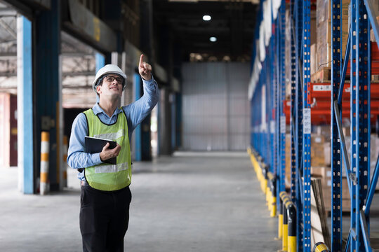 Caucasian male warehouse worker wearing white hard hat and safety vest standing in warehouse while using digital tablet manage inventory, organize logistics operations in distribution center