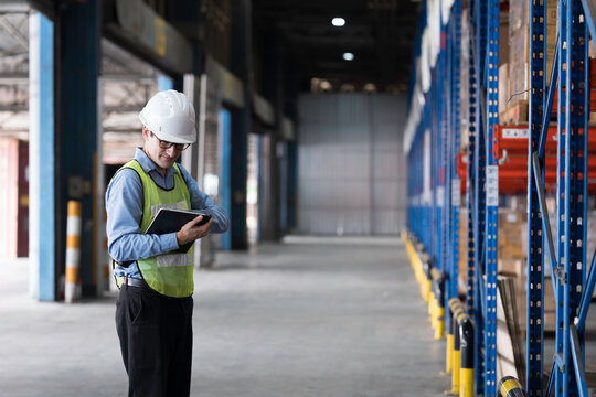 Caucasian male warehouse worker wearing white hard hat and safety vest standing in warehouse while using digital tablet manage inventory, organize logistics operations in distribution center