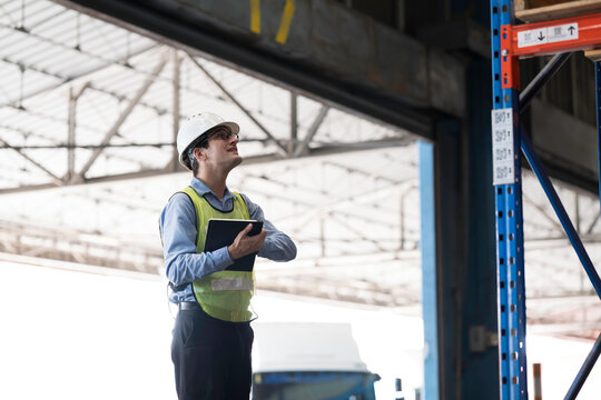 Caucasian male warehouse worker wearing white hard hat and safety vest standing in warehouse while using digital tablet manage inventory, organize logistics operations in distribution center