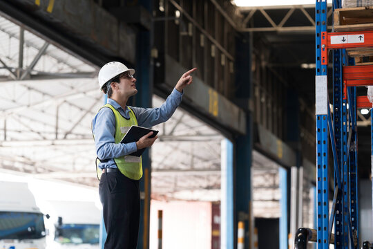 Caucasian male warehouse worker wearing white hard hat and safety vest standing in warehouse while using digital tablet manage inventory, organize logistics operations in distribution center