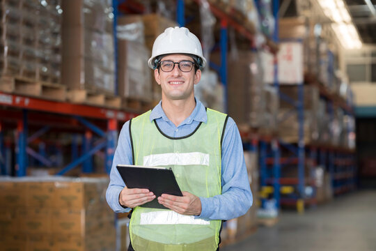 Male warehouse worker wearing white hard hat and safety vest standing in warehouse while holding digital tablet for inventory management in large distribution center