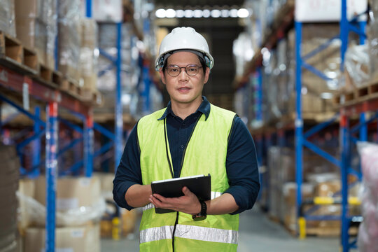 Male warehouse worker wearing white hard hat and safety vest standing in warehouse while holding digital tablet and checking inventory stock with high shelves and cardboard boxes in background