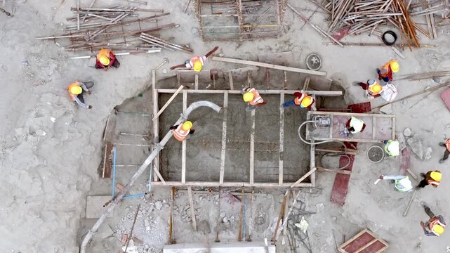 Top-down drone shot of workers pouring ready mix concrete into wooden shuttering for an RCC foundation footing at a construction site in India using a concrete pump. 