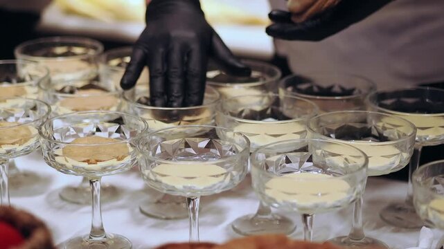 Caterer fills rows of dessert glasses with cream at a serving table