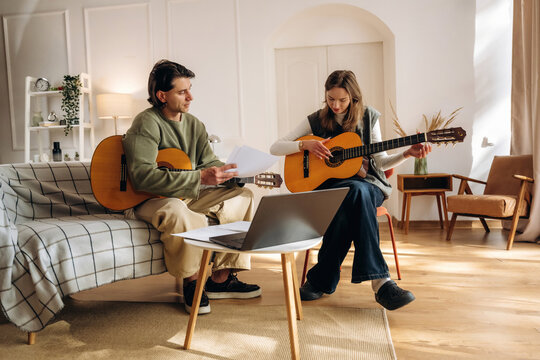 Two people playing acoustic guitars together in a bright modern living room with a laptop, sheet music, sofa, and natural light indoors