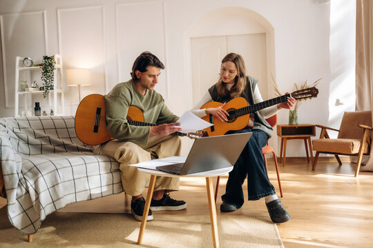 Man and woman playing acoustic guitars together while reading sheet music in a bright living room with laptop and modern home interior