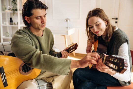 Man teaching woman to play acoustic guitar while sitting together in a bright home interior, relaxed musical lesson and shared learning moment indoors