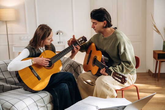 Two people playing acoustic guitars together in a bright home interior with sheet music and seating, musical practice scene