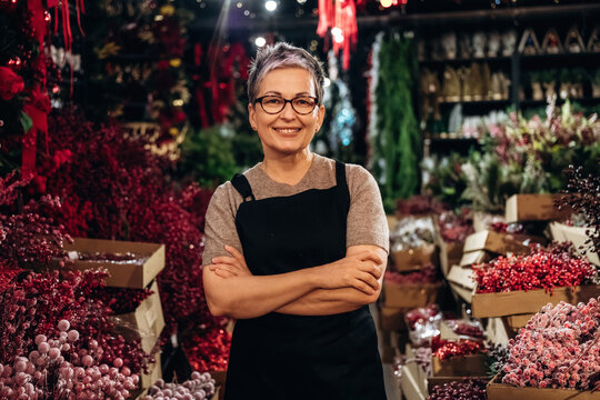 Smiling mature woman with crossed arms wearing an apron among colorful floral arrangements and plants inside a flower shop interior
