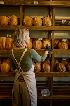 Woman organizes artisan bread on wooden shelves in a warm bakery