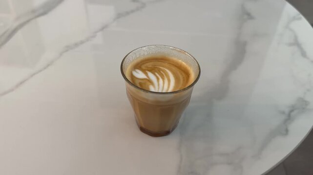 A small glass of milk coffee with a white rosetta-style latte art pattern sits on a white and grey marble surface.