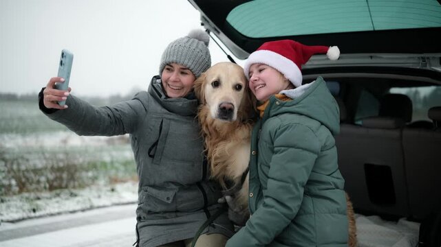 Happy Mother And Daughter Wearing Santa Hats Take A Selfie With Dog In A Car Trunk During A Winter Road Rest Stop