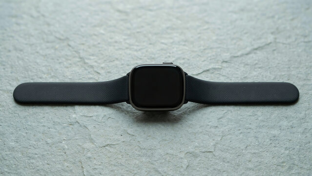 A black smartwatch with a black silicone strap lying flat on a textured grey stone surface, down view of a wearable technology device with a dark screen