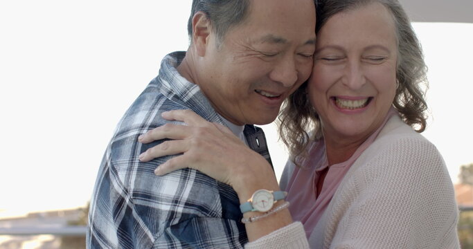 Senior couple standing on balcony by water embracing with wristwatch, bracelet and clip on mic