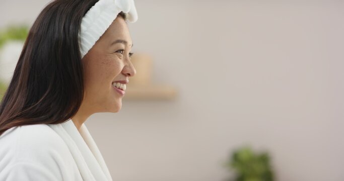 Woman wearing white robe with towel headband, smiling in bathroom with wooden shelf and green plant