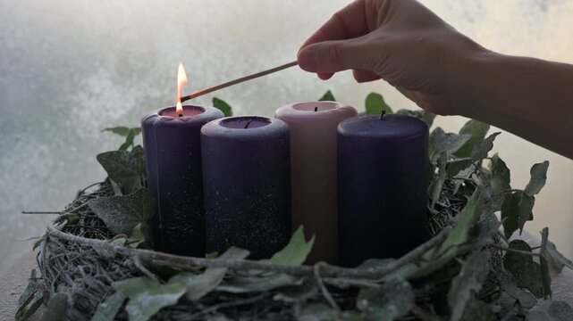 Hand lighting Advent candles candles set three purple candles and one pink in a rustic wreath by frosty window.