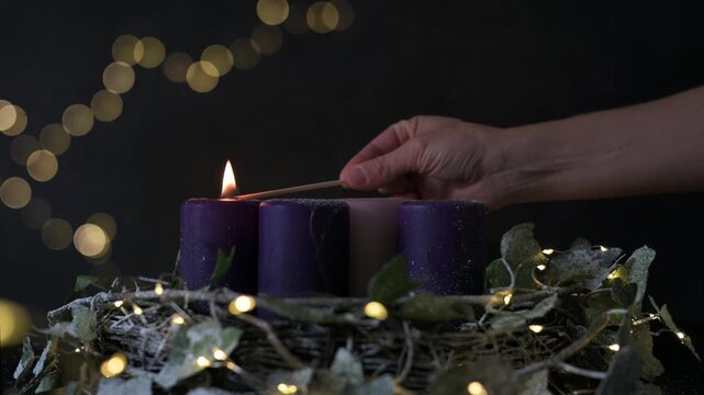 A hand lighting candles on a Advent wreath. Features purple and pink pillar candles in the dark. Ideal for religious, spiritual, and holiday-themed video content.