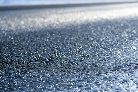 Transparent black ice road surface showing glossy frozen water droplets, fine fissures, coarse asphalt texture, bright winter sunlight, reflective pavement, and dangerous driving conditions