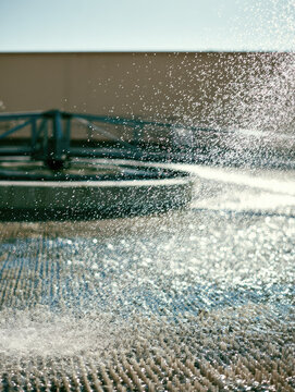 Water spray over trickling filter packing media municipal treatment facility, sunlight creating sparkling droplets above textured infrastructure with industrial wall background and calm atmosphere
