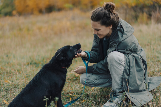 Smiling young woman in a raincoat gently holding her black dog's face in her hands during a walk on an overcast autumn day.