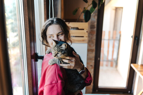 Close up portrait of young woman holding cat and looking out window together