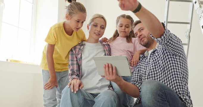 Parents daughters family tablet home renovation planning. Father points at a screen while family reviews decorating plans in a bright sunlit unfinished room. A vision for family living.