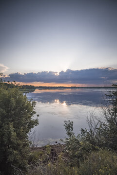 An evening lakeside scene, with the sun shining from behind the clouds and reflecting off the water&mdash;a warm landscape captured just before sunset