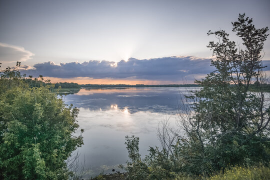 An evening lakeside scene, with the sun shining from behind the clouds and reflecting off the water&mdash;a warm landscape captured just before sunset