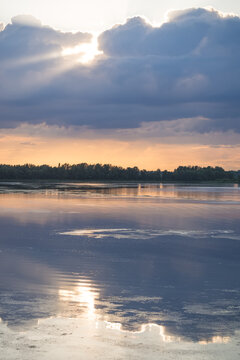An evening lakeside scene, with the sun shining from behind the clouds and reflecting off the water&mdash;a warm landscape captured just before sunset