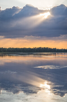 An evening lakeside scene, with the sun shining from behind the clouds and reflecting off the water&mdash;a warm landscape captured just before sunset