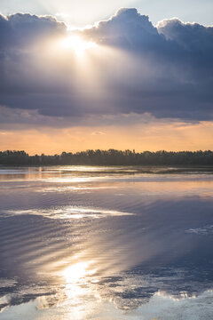 An evening lakeside scene, with the sun shining from behind the clouds and reflecting off the water&mdash;a warm landscape captured just before sunset