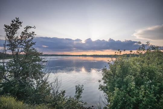 An evening lakeside scene, with the sun shining from behind the clouds and reflecting off the water&mdash;a warm landscape captured just before sunset