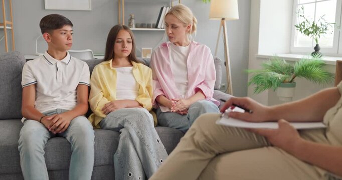 Mother and teenagers attend family counseling together. A psychologist takes notes as they discuss important feelings during a private therapy consultation. A supportive family care concept.