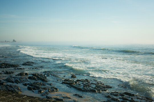 Rocky ocean coastline with breaking waves and distant buildings fading into coastal haze