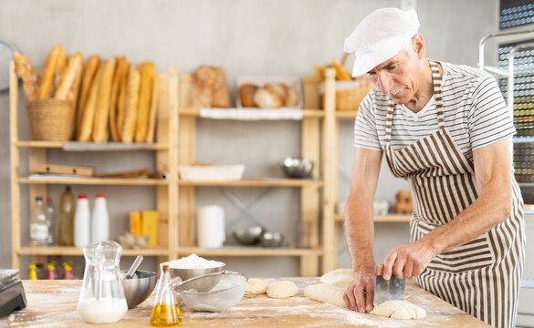 Mature baker uses knife to cut pieces of raw dough to make croissants