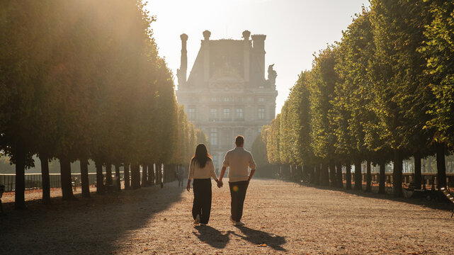 A couple walks along a broad tree-lined avenue in Paris, France, as warm late afternoon light casts long shadows toward an elegant historic building. Jardin des Tuileries