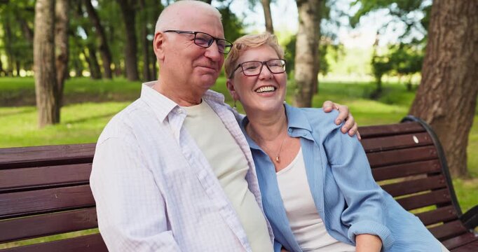 Seniors couple laughing embracing on park bench. Happy partners relax outdoors, sharing warmth in a leafy park. Genuine closeness and happiness. Strong concept of joyful retirement love.