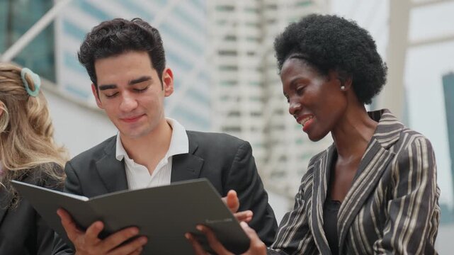 Two professionals converse in an urban setting. Man holds a folder, looking at the woman. She listens attentively, smiling slightly