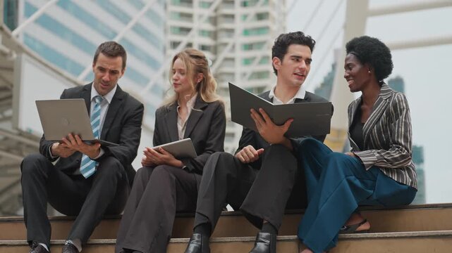 Four professionals in business attire sit on a modern city ledge. They use laptops, tablets, and folders for work