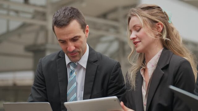 Two professionals collaborate outdoors near a building. They examine digital devices with focused expressions. The man speaks