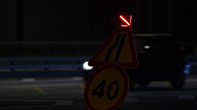 Temporary road narrowing warning sign with 40 kmh speed limit and illuminated directional arrow at night, showing traffic safety measures and road maintenance with passing vehicles