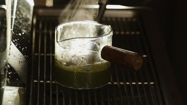 Light steam flow rises over fresh matcha in espresso measuring glass closeup. Hot green tea made of plant powder on coffee maker tray