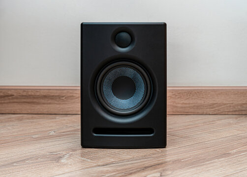 A sleek black speaker is positioned on a wooden floor, showcasing its modern design. The speaker's circular elements and smooth finish highlight its aesthetic appeal.