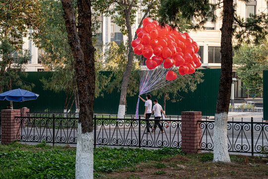 Two individuals walk through a park, carrying a large cluster of red balloons. The vibrant balloons contrast with the greenery and urban setting, creating a lively scene.