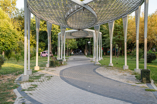 A scenic park pathway features a beautifully designed pergola structure with intricate patterns. The walkway is surrounded by lush greenery, creating a serene atmosphere.