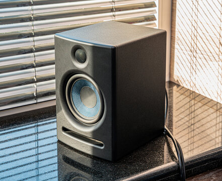 A sleek black speaker sits on a sunlit surface, casting shadows through nearby blinds. The speaker's design is modern, enhancing its appeal in a contemporary setting.