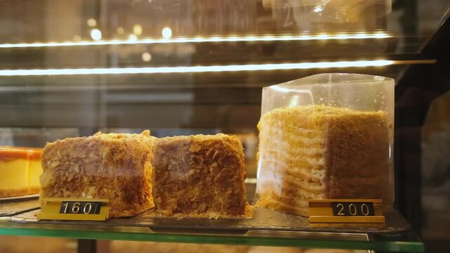 Layered cake pieces sit inside glass bakery display. Dessert slices show crisp pastry texture near price tags under warm cafe lights