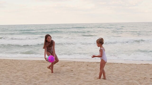 Two young girls wearing swimsuits play catch with a bright ball on a sandy shore by the sea