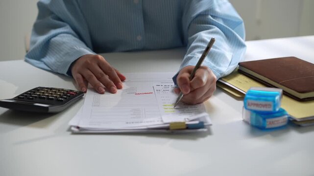 Office worker reviewing paperwork, completing form with calculator and correction tape desk, carefully checking financial document details while writing notes with pen during routine administrative