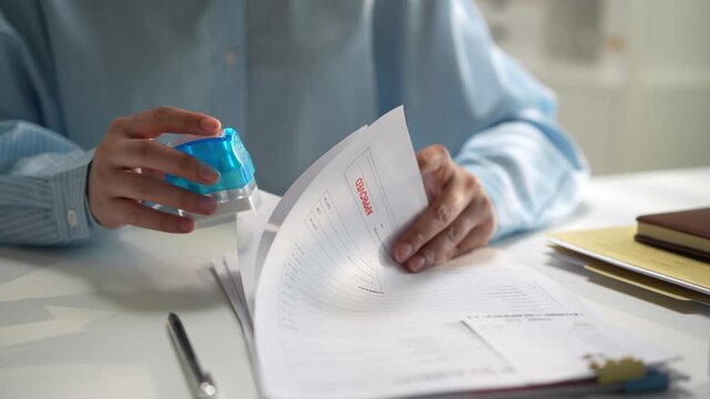 Businessperson reviewing document at office desk, turning page and handling paper form with hand, pen, blue stamp, and highlighter for careful paperwork review and focused attention